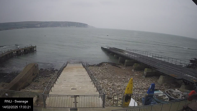 A coastal view showing a concrete ramp leading down to a rocky shoreline and the sea. On the left, there is a wooden pier extending into the water. On the right, a metal ramp leads to a small, partially submerged dock. Several colorful kayaks are stacked near the ramp, and the scene is set against a grey sky with low visibility. The water appears calm with small waves.