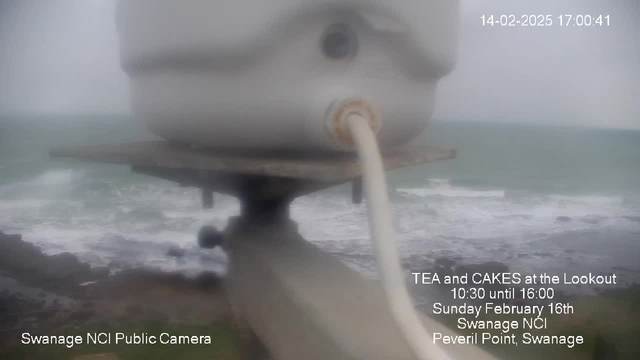 A blurred view from a webcam, displaying a coastline with waves crashing against rocky shores. The foreground shows part of the camera structure, with a visible cable. The sky is overcast, indicating possible rainy weather. Text overlays at the bottom mention a public event (tea and cakes) at Swanage NCI on February 16th.