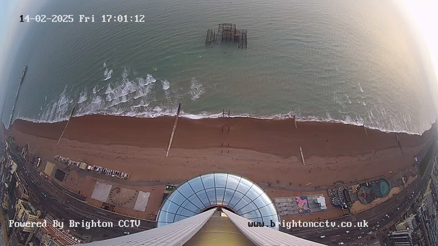 A view from a high vantage point looking down at a beach with light brown sand. The ocean is visible, with gentle waves lapping at the shore. There are two structures protruding from the water: one is a pier on the left and the other is a partially submerged, old pier in the center. The beach is lined with a road and several buildings, including a circus-like structure with a colorful design. It is dusk, and the sky shows a gradient of colors. The timestamp on the image indicates it is February 14, 2025, at 17:01:12.