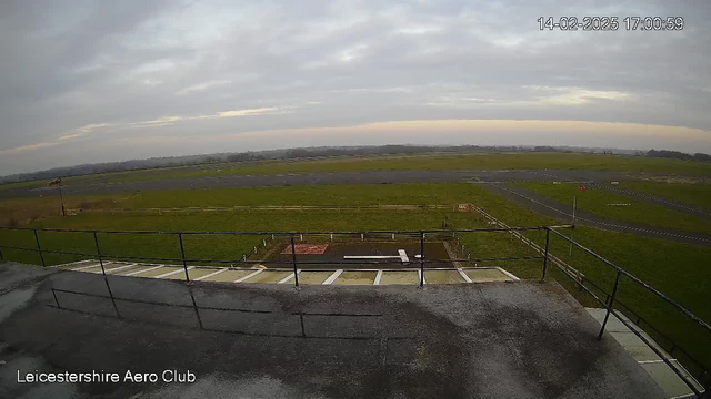 A view from a webcam overlooking a grassy area and runway. In the foreground, there are concrete steps leading down from a building, with a railing along the edge. To the left, a windsock is visible, indicating wind direction. In the distance, a gray, overcast sky looms above, while the horizon reveals rolling hills. The area features a mix of green grass and gravel, with a clear runway running horizontally across the image. There are some markers and lines on the runway, and a few signs are positioned nearby.