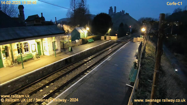 A railway station platform is shown in dim evening light. On the left, there is a stone building with multiple windows and a green bench. To the right, there are two parallel railway tracks and more benches along the platform. In the background, a hill rises, featuring castle ruins. Additional lighting fixtures are present along the platform and on the adjacent path, where a sign reading "Way Out" is visible. The scene conveys a quiet, tranquil atmosphere typical of rural stations.