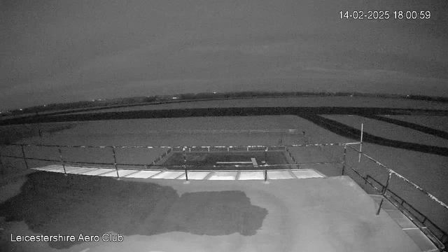 A black and white webcam image showing a wide view of an empty, flat field under a cloudy sky. The foreground features a wooden platform with a railing, and a small area with a rectangular shape likely indicating a part of a structure. The background features a subtle horizon line with some distant lights possibly suggesting buildings or vehicles. The overall scene conveys a quiet, deserted atmosphere. The timestamp on the image reads 14th February 2025 at 18:00:59.