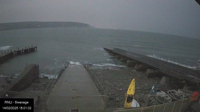 A view of a rocky shoreline at dusk, with a pier extending into a choppy sea. The sky is overcast, and the water appears grayish-green. In the foreground, there is a concrete pathway leading to the pier. To the right, two kayaks, one yellow and one blue, are stored near the edge of the pathway. Waves are lapping against the rocks, and a distant landmass is visible across the water.