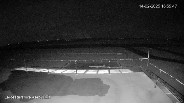 A nighttime webcam view of the Leicestershire Aero Club. The image is mostly dark, with silhouettes of a roof and railing visible. There are faint lights in the distance, hinting at a surrounding landscape. The timestamp in the corner reads 18:59:47 on February 14, 2025.