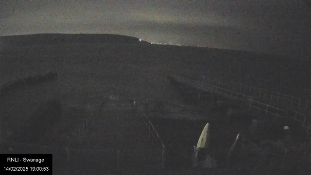 A dark scene showing a landscape with very low visibility. There is a faint outline of a hill in the background, and some lights are visible at a distance. In the foreground, there are silhouettes of boats, with one boat partially illuminated. There is a wooden structure or pier running horizontally across the lower part of the image. The overall atmosphere appears quiet and serene, with cloudy skies above.