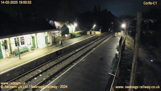 A dimly lit railway station at night. The platform features a stone building with large windows on the left, a green bench, and a sign that reads "WAY OUT." There are wooden barriers at the far end of the platform, with additional benches and a light illuminating the area. The tracks run alongside the platform, which appears quiet with no visible trains. In the background, some trees and a faintly lit house can be seen.