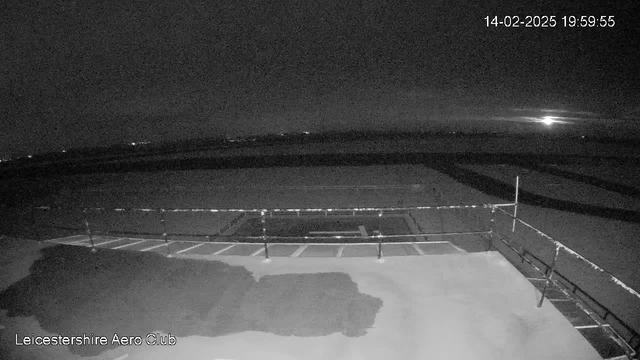 A night view of an airfield at Leicestershire Aero Club. The image is mostly dark, with faint outlines of a hangar and a runway. A fence is visible in the foreground, with a few horizontal bars. In the distance, there is a faint light or glow on the horizon, indicating either streetlights or a rising moon. The timestamp shows the date and time as February 14, 2025, at 19:59:55.