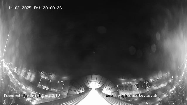 A black and white webcam image shows a view from a high vantage point looking downwards. The lower half features a wide, bright light leading towards an unknown area, with visible lines suggesting a structure below. In the upper half, the sky is dark with some indistinct shapes and blurred spots, possibly caused by moisture or lens distortion. A timestamp at the top indicates it is February 14, 2025, at 20:00:26. The bottom left corner displays "Powered By Brighton CCTV."