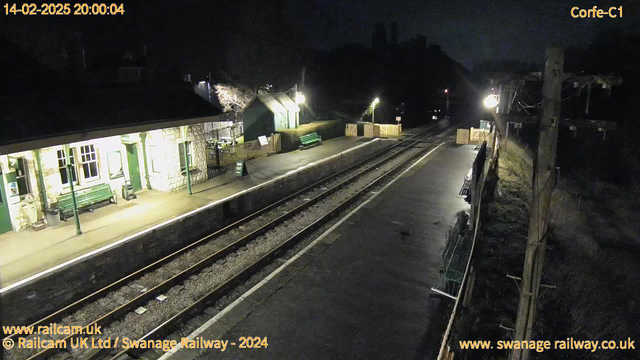 A dimly lit train station at night, with stone walls and green benches along the platform. A small building on the left has several windows, while a slight hill can be seen in the background. To the right, a wooden fence leads to an exit, and there are two green benches visible. The tracks are empty, surrounded by gravel, and faint lights illuminate the scene, reflecting off the wet pavement.