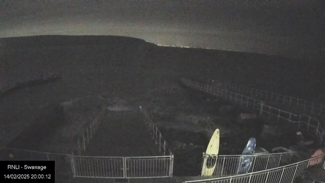 A dark, mostly shadowy scene depicting a coastal area at night. There are two kayaks resting on the ground: one yellow and one blue. In the background, faint outlines of a hill or land can be seen against a slightly lighter sky. A wooden walkway or path is visible, bordered by metal railings. Overall, the image has low visibility with minimal light, creating a nighttime atmosphere.