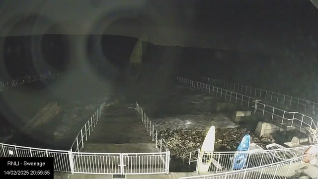 A dark scene of a beach area at night. In the foreground, there is a wooden pier extending into the water, surrounded by a white railing. To the right, there are two surfboards standing on the ground, one yellow and one blue. The water appears choppy with noticeable waves. In the distance, faint lights appear along the horizon, indicating a nearby shoreline or town. The overall atmosphere is calm yet somewhat eerie due to the darkness.