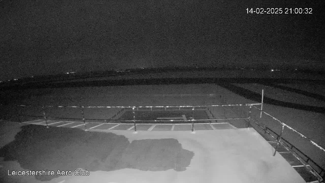 A dark, nighttime scene showing a large, flat surface that appears to be an airstrip. In the foreground, there is a railing or fence bordering the area. The lighting is low, with a few distant lights visible in the background, indicating a few structures or possibly other vehicles. The overall atmosphere is quiet and serene, typical of a late evening at an aerodrome. The timestamp in the upper right corner displays the date and time.