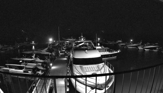 A nighttime image of a marina, featuring several boats docked along a wooden pier. The scene is illuminated by bright lights reflecting off the water. The majority of the image is dark, with the silhouettes of boats visible against the night sky. Some boats are larger yachts, while others appear to be smaller vessels. The perspective is from above the dock, looking down the row of boats.