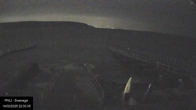 A dimly lit scene by the water at night. The silhouette of a pier extends into the distance, partially illuminated by faint lights on the horizon. To the foreground, several boats are visible, with one boat featuring a white sail. The surroundings appear calm and quiet, with a dark landscape in the background. A timestamp indicating the date and time is present in the lower left corner.