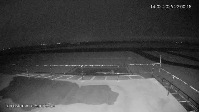 A dark, nighttime scene from a webcam at Leicester Aero Club. The image shows a flat, empty surface, likely an airstrip or runway, under low light conditions. In the foreground, there is a railing or fence. The background features a horizon with faint lights, suggesting distant structures or vehicles. The date and time are displayed in the top right corner.