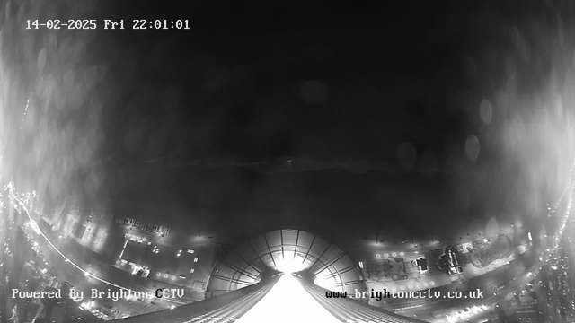 A black and white image from a webcam showing a cityscape at night. The upper part is dark, while the lower section features blurred lights from vehicles and buildings. There is a bright area in the center, likely reflecting light, and many circular shapes indicating out-of-focus lights. The time and date are displayed in the upper left corner, while the source information appears at the bottom.