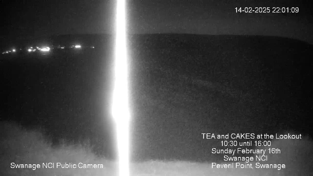 A dark webcam image showing a nighttime view of a landscape with faint lights in the distance, possibly representing a shoreline or buildings. The foreground is mostly shadowed, while a bright vertical line runs through the center, likely a reflection or lens flare. Text in the lower portion displays event information and identifies the camera location as Swanage NCI Public Camera. The date and time are noted at the top right corner.