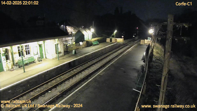 A dimly lit train station at night, featuring two platforms. On the left, there is a stone building with large windows, a green bench, and a sign reading "Way Out." The right side shows another green bench, a section of the platform, and wooden fencing. There are railway tracks running between the platforms, with gravel visible. In the background, there are hints of trees and a distant light source. Overall, the scene is quiet and mostly empty.