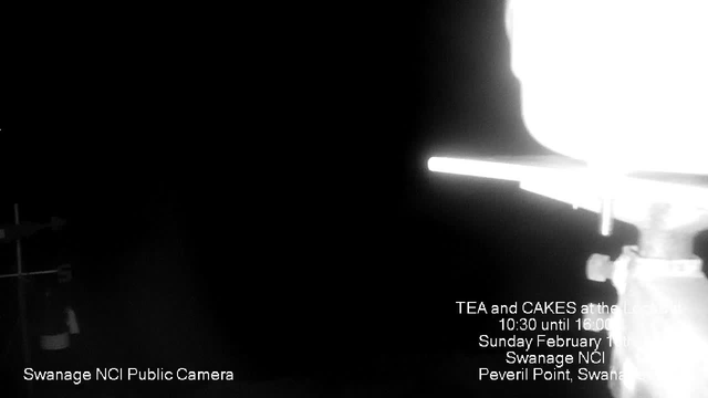 A dark image featuring a partially obscured sign with white text indicating a public camera location at Swanage NCI. The lower part includes details about a tea and cake event on February 11th, mentioning the time and place, with the words "Swanage NCI, Peveril Point, Swanage" visible. There are faint outlines of objects possibly related to the camera setup on the left side.