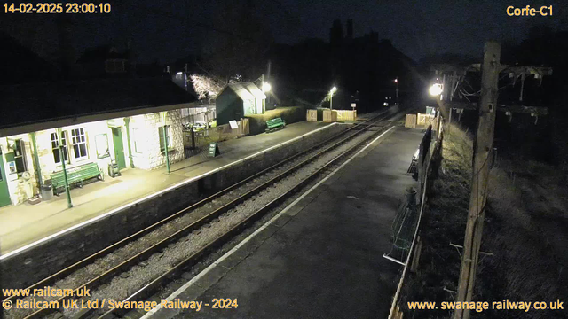 A nighttime view of Corfe Castle railway station. The platform is mostly empty, with a few benches and a sign indicating the way out. The station building, built from light-colored stone, has large windows and is illuminated by lights. The railway tracks run alongside the platform, extending out of view. In the background, low hills and tree silhouettes are visible against the dark sky.