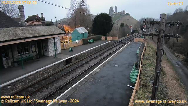 A railway station scene at dawn, showing an empty platform with stone and metal benches. On the left, there's a building with a sloping roof and a green door, partially obscured by trees. In the background, a grassy hill rises up, featuring an ancient stone castle. The railway tracks extend towards the horizon, with a signal post displaying red lights. A wooden fence encloses some colorful decorations and a sign indicating "WAY OUT." The sky is light grey, suggesting early morning.