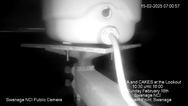 A close-up image of a white container, possibly a jug, placed on a metal platform. The container has a hose or cable connected to it, leading down towards the platform. The background appears dark, and there is text overlaying the image that includes event details: "Tea and Cakes at the Lookout, Sunday February 16th, Swanage NCI, Peveril Point, Swanage." The time stamp on the image indicates it was taken on February 15, 2025, at 7:00 AM.