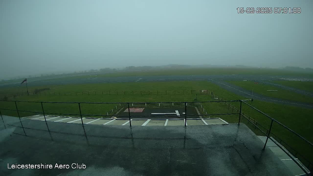 A view from a webcam at the Leicestershire Aero Club shows an open field under cloudy and foggy conditions. The ground is wet, possibly indicating recent rain. In the foreground, there are railings and a flat surface, likely a deck or observation area. The background features a grassy field with a runup area and some faint outlines of runway markings. Visibility appears very low due to fog, with no distinct landmarks visible beyond the immediate vicinity. A flag is seen on a pole to the left of the frame. The time shown on the image is 07:01 on February 15, 2025.