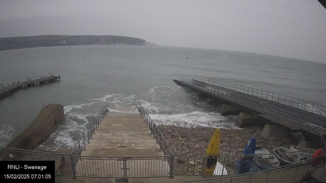 A view of a coastal area with a gray sky and calm sea waters. In the foreground, there are steps leading down to the shoreline, surrounded by a railing. To the left, there is a small pier extending into the water. Several colorful kayaks are positioned near the edge, one yellow and one blue. The beach area consists of pebbles and rocks, while a distant cliff with greenery can be seen in the background. The overall scene has a tranquil yet slightly overcast atmosphere.