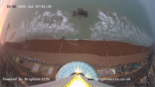 A high-angle view of a beach at dawn, with waves gently crashing onto a sandy shore. In the distance, a broken pier extends into the water. The foreground features part of a tall structure, with a glass dome reflecting the early morning light. The beach area is mostly empty, with a few structures and a winding promenade visible to the right. The date and time are displayed at the top left corner of the image.