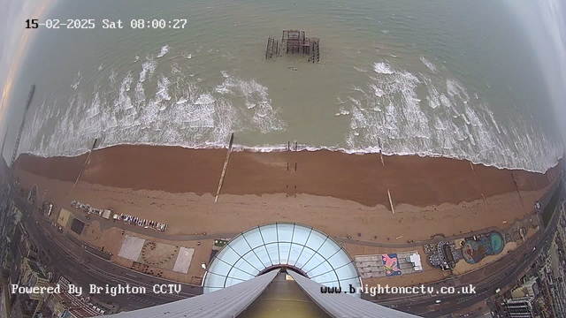 Aerial view of a beach with dark sand and light blue ocean waves. The image shows a pier extending into the water. A partially visible, dilapidated structure lies in the water near the pier. In the bottom part of the frame, there is a circular glass structure, likely a building, with several lines suggesting a height, indicating the perspective is from above. Along the shoreline, there are some buildings, a visible amusement park, and a road running parallel to the beach. The sky appears cloudy, and the time displayed is 08:00:27 on 15 February 2025.