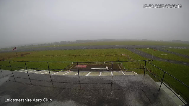 A foggy view of an airfield taken from a balcony. The scene shows a grassy area and an empty runway with various markings. A red flag is visible on a pole to the left, indicating wind direction. The atmosphere appears overcast, with limited visibility into the distance. The lower part of the image features a railing and steps leading down from the balcony.