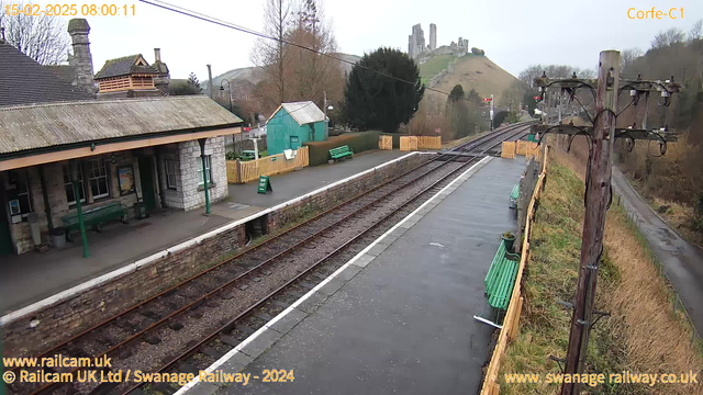 A view of Corfe Castle station on a cloudy day. The foreground features a stone platform with several green benches and a small green shed. A few trees are located on the left side, and a wooden fence runs along a path. In the background, Corfe Castle ruins are visible atop a hill, with railway tracks extending from the left side of the image into the distance. The scene has a generally gray and muted color palette.