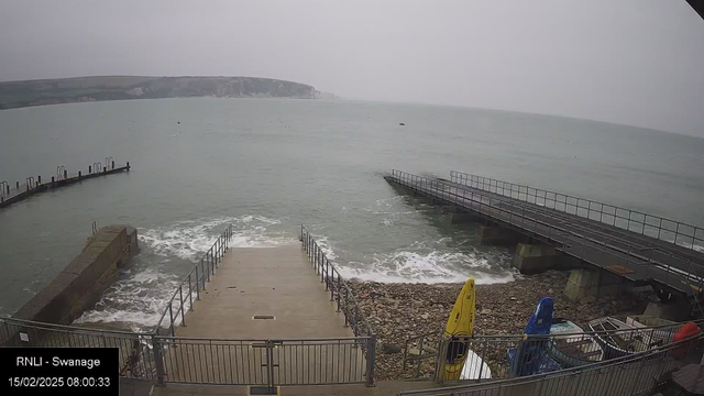 A cloudy scene at a coastal location. In the foreground, there is a set of concrete steps leading down to the water, surrounded by metal railings. Waves are gently crashing at the base of the steps. To the right, a long wooden pier extends into the water, with a few boats and kayaks stacked beside it. The background features a grey, rocky coastline and rolling hills under an overcast sky.