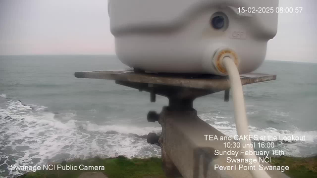 A webcam image shows a cloudy sky above a coastline with waves crashing against rocks. In the foreground, there is a white container mounted on a stand, with a pipe extending from it. The text in the lower left corner indicates the camera is from Swanage NCI Public Camera. Additional text on the image promotes a tea and cake event on February 16th at Peveril Point, Swanage, from 10:30 to 16:00.