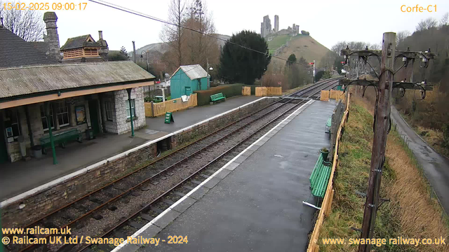 A railway station scene with a stone station building featuring a peaked roof, green benches, and a small blue shed. In the background, there are green hills with the ruins of a castle on top. The tracks, which are visible in the foreground, run parallel to a paved path. Surrounding vegetation is sparse with some hedges and a wooden fence. The weather appears overcast, and the ground is wet, likely from recent rain. A sign reads "WAY OUT."