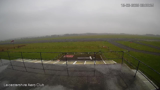 A cloudy and overcast scene at the Leicestershire Aero Club. In the foreground, there is a railing with a view down to a grassy area and a tarmac runway. The runway stretches across the middle of the image, showing a wide, empty space. A small building or shelter is visible on the ground level. In the distance, there are blurred outlines of trees and land, emphasizing a gloomy, foggy atmosphere. A wind sock is seen, indicating the wind direction, but it is not billowing strongly due to the calm conditions.