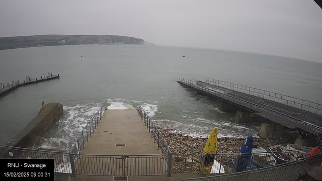 A coastal scene shows a gray sky over the sea. In the foreground, there is a set of stairs leading down to the water, with waves splashing against the steps. To the right, a rocky area is visible, with a couple of colorful kayaks, one yellow and one blue, positioned near the quay. In the distance, the shoreline features cliffs, slightly blurred due to the overcast weather. A pier extends into the water, with several posts visible along its edge.