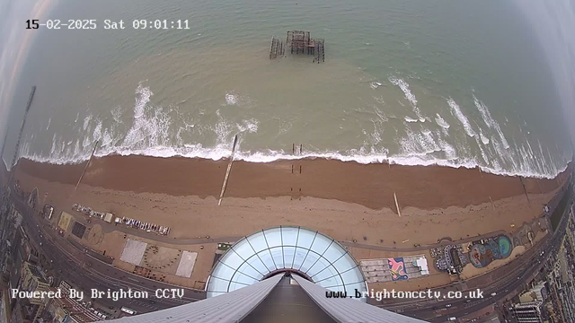 Aerial view of a beach with brown sand and waves gently crashing. In the foreground, the base of a large observation tower is visible, leading to a glass dome. The shoreline features a pier extending into the sea, with a structure partially submerged in the water. The background shows a cloudy sky and a wide stretch of ocean. There are some buildings and colorful structures along the beach.