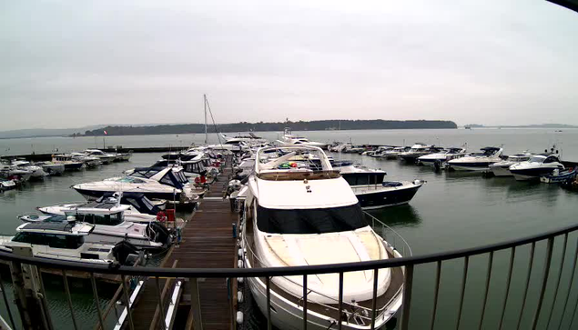 A marina scene with numerous boats docked in calm water. In the foreground, a large white motorboat is positioned beside a wooden walkway. Surrounding it are various smaller boats, some with blue and white striped coverings. The sky is overcast, and the distant shoreline is visible, along with a hint of greenery. The atmosphere appears tranquil, with no visible people in the image.