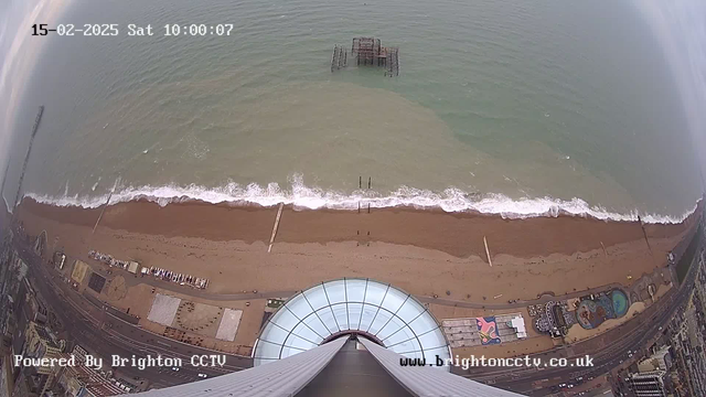 Aerial view of a sandy beach with gentle waves lapping at the shore. In the foreground, the top of a large circular structure is visible, likely a viewing platform. Below, several lines of wooden posts are partially submerged in the water, and an old pier can be seen extending from the shore. The beach features a few scattered structures and colorful patterns on the ground nearby. To the left, there is a section of coastal road with vehicles, and the sky is partly cloudy.