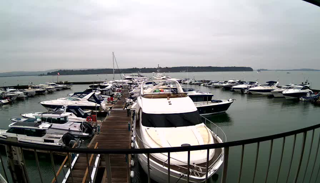 A view of a marina filled with several boats and yachts of varying sizes moored along a wooden dock. The sky is overcast with a gray hue, reflecting on the calm water. In the background, additional boats can be seen further out on the water, with trees lining the shore in the distance. A railing is visible in the foreground, framing the scene.