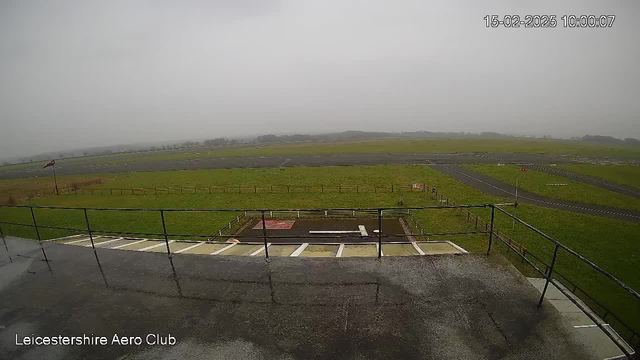 A view from a webcam at the Leicester Aeroclub showing a wide expanse of grassy field and a runway. The scene appears overcast and misty, with low visibility. In the foreground, there is a railing and steps leading down to the ground. The background features a runway with markings and a few signs. The sky is gray, indicating a cloudy atmosphere.