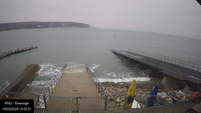 A cloudy view of a coastline with calm water. In the foreground, a concrete ramp leads down to the water, lined with metal railings. To the left, there is a rocky shoreline and a small pier extending into the sea. Several colored kayaks are stored to the right, with a bright yellow one prominent. The background features green hills and cliffs rising above the waterline. Time stamp indicates the date and time of the image.