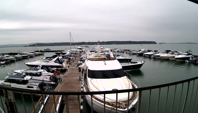 A view of a marina filled with various boats and yachts docked at wooden piers. The scene is set under a cloudy sky, with the water calm, reflecting the grey tones of the sky. Several boats are lined up in the foreground, with some people visible near the dock. In the background, a few boats are anchored farther out on the water, and distant land can be seen along the horizon.