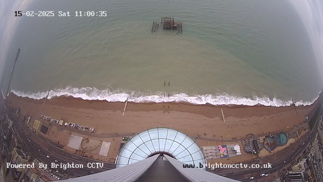 Aerial view of a beach along the coastline, featuring sandy shores and gentle waves. The scene includes an abandoned pier structure in the water and various beach amenities scattered along the sand. A circular glass structure at the bottom indicates a building or tower. The sky is overcast with clouds, and the time and date are displayed at the top of the image.