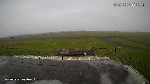 A cloudy, overcast sky is visible above a grassy airfield. In the foreground, a flat rooftop railing is seen, alongside several white and gray stripes on the surface. Below, the airfield stretches out with green grass, a gray runway in the center, and scattered trees in the distance. A windsock stands at the far left, indicating wind direction. The image has a timestamp indicating it was taken at 11:00 on February 15, 2025, at Leicestershire Aero Club.