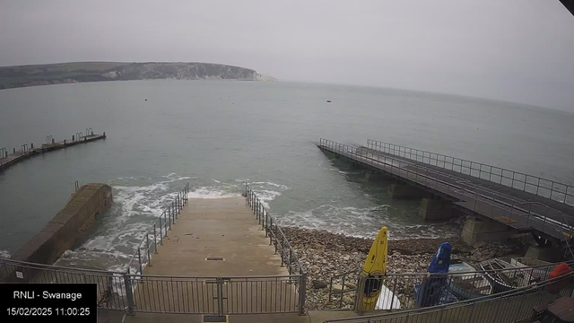 A gray overcast sky looms above a coastal scene. In the foreground, concrete steps lead down to the water, with waves gently lapping at the base. To the left, a small stone breakwater juts out into the sea, while a jetty extends further out to the right. Several colorful kayaks are visible on the shore near the steps. The sea appears calm, with a distant shoreline of cliffs in the background.