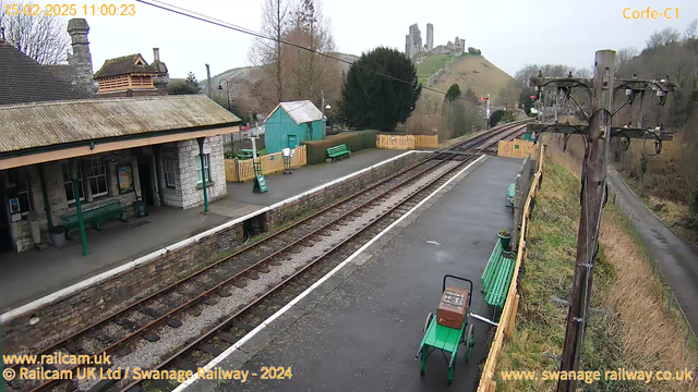 A view of Corfe Castle railway station on a cloudy day. The image shows a platform with green benches and a vintage cart. To the left, there is a stone building with a sloped roof and multiple windows. In the background, the silhouette of Corfe Castle can be seen atop a hill. The railway tracks run through the middle of the image, leading towards the hills in the distance. There are trees and a small green shed visible nearby, adding to the rural atmosphere.