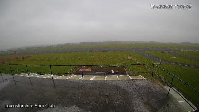 A view from a webcam showing a cloudy, overcast sky. In the foreground, there is a railing along a viewing deck, with a wet surface suggesting recent rain. Below the deck, there is an area with a gray surface, possibly a landing area, bordered by green grass. In the distance, there are several grassy fields and a faint horizon line. A small red flag can be seen to the left, indicating the presence of wind. The overall scene appears serene and quiet, reflecting a calm day at the airfield.