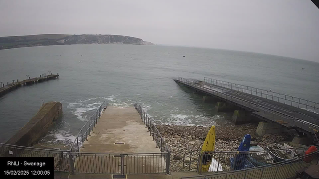 A coastal scene featuring calm waters under a gray sky. In the foreground, a concrete ramp descends toward the water, flanked by a metal railing. To the right, a wooden pier extends into the sea, with another smaller pier to the left. On the ground, there are colorful kayaks: one yellow and one blue. The shoreline is rocky with some scattered stones visible, and in the distance, cliffs are faintly visible along the horizon.